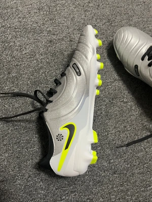 Nike Tiempo grå fotbollsskor - Säljer ett par Nike Tiempo fotbollsskor i grått med svarta och neongula detaljer. Skorna har snörning, rund tå och platt sula med dobbar för gräsplan. Ovandelen är i syntetmaterial och insidan har en fräsch neongul färg. Perfekta för dig som vill sticka ut på planen.