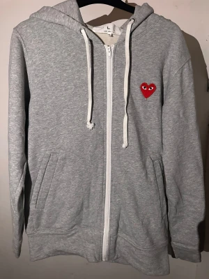 Grå ziphoodie från Comme des Garçons Play - Grå zip-hoodie från Comme des Garçons Play med vit dragkedja och snören. I en värdigt fin skick, i storlek L/M. Priset kan diskuteras men tyvärr inte mycket då tröjan är i väldigt fin skick!