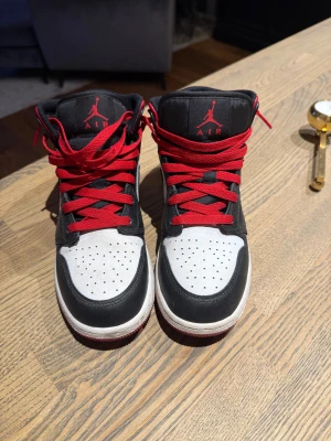 Nike Air Jordan 1 Mid svart/vit/röd - Nike Air Jordan 1 Mid sneakers i svart, vit och röd med klassisk swoosh på sidan och röda snören. Skorna har perforerad tå, högt skaft och ikonisk Jumpman-logga på plösen. Materialet är skinn och syntet, med platt sula och rund tå. Perfekt för dig som gillar streetstyle.