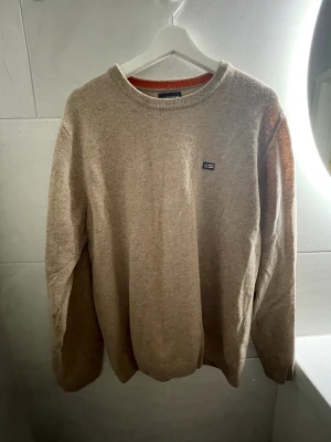 Beige stickad tröja från Lexington - Snygg beige stickad tröja från Lexington med rund hals och diskret logga på bröstet. Insidan av kragen har en cool orange ribbad detalj som ger extra stil. Perfekt för dig som gillar klassisk men ändå chill look.