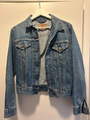 Blå jeansjacka från Levi's,  - Snygg klassisk blå jeansjacka från Levi's i storlek S. Jackan har bronsfärgade knappar framtill och på fickorna, två bröstfickor med lock och lång ärm. 