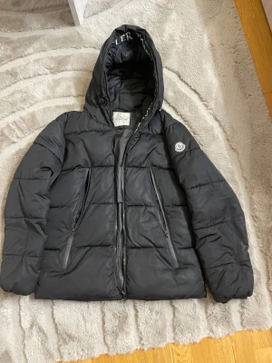 Svart Moncler dunjacka med huva - Svart moncler dunjacka med stor huva och logotyp på ärmen. Jackan har två snedställda fickor med dragkedja och quiltad design. Insidan av huvan har Moncler-tryck och ett färgglatt serietryck finns på fodret. Perfekt för kalla dagar och riktigt snygg streetstil. Kan diskutera pris. DRAGKEDJAN ÄR TRASIG OCH DEN ÄR INTE ÄKTA! 