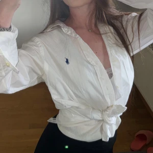 Vit skjorta från Ralph Lauren - Vit skjorta från Ralph Lauren med klassisk krage och blå broderad logga på bröstet. Skjortan är knäppt framtill och har långa ärmar samt är knuten i midjan för en trendig look. Tillverkad i bomull och passar perfekt till jeans eller kjol.