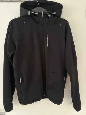 Svart vindjacka Peak Performance S - Svart vindjacka från Peak Performance med huva och dragsko, hel dragkedja framtill och två fickor med dragkedja. Jackan har en stilren look med diskret logga på bröstet och bak på huvan. Perfekt för dig som gillar sportig och clean stil.