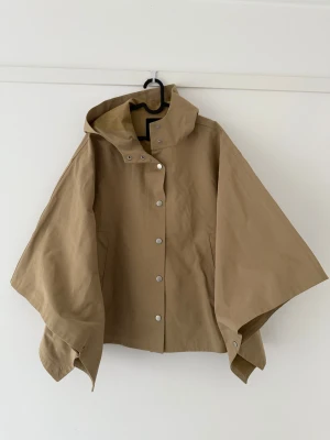 Beige regnjacka i cape/poncho-stil från Zara - Beige regnjacka från Zara i poncho/cape-modell med huva och tryckknappar framtill. Jackan har vida ärmar och en loose passform, perfekt för regniga dagar. Tillverkad i ett slätt syntetmaterial som står emot väta och har minimalistisk design. Jackan har en längd på ca 68 cm. Viral, trendig, och slutsåld!!