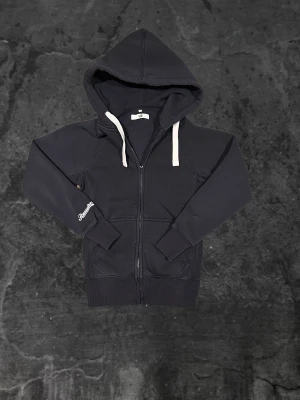 Russemerch zip hoddie  - Svart hoodie från Russemerch i storlek XS med vit snörning och dragkedja framtill. Tröjan har huva, ribbade muddar och fickor på sidorna. Broderad logga på ärmen ger en sportig vibe. Perfekt för chill eller häng med kompisar.