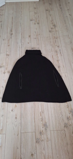 Svart stickad poncho från Joseph - Svart stickad poncho från Joseph med hög krage och två diskreta fickor framtill. Den har en loose passform och är tillverkad i ett mjukt, grovstickat material som passar perfekt för lager-på-lager-stil. Camel hair