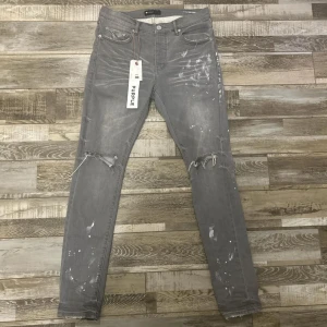 Grå skinny jeans från Purple - Snygga grå jeans från Purple med skinny passform och slitna detaljer vid knäna. Jeansen har coola färgstänk och trasiga partier som ger en edgy vibe. Klassisk femficksmodell med knapp och dragkedja framtill. Size 32. Fraktar inom 24 timmar.
