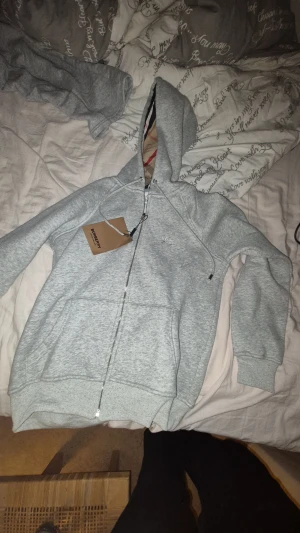 Grå hoodie från Burberry med dragkedja xxs - Snygg ljusgrå hoodie från Burberry i storlek m men sitter som xxs. Tröjan har huva med snörning, hel dragkedja och broderad Burberry-logga på bröstet. Tillverkad i mjuk bomullsmix med ribbade muddar och fickor framtill. Perfekt för chill dagar.