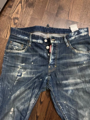 Dsquared2 blå jeans med slitningar - Snygga blå jeans från Dsquared2 med coola slitningar, färgstänk och patchade detaljer. Jeansen har klassisk femficksmodell, knappgylf och tydliga kontrastsömmar. Perfekt för dig som gillar en streetig och avslappnad look.