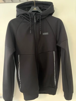 Svart vindjacka från McKenzie med huva - Svart vindjacka från McKenzie med hel dragkedja, huva med snörning och diskreta grå detaljer längs fickorna. Jackan har en clean look och är tillverkad i ett mjukt syntetmaterial. Perfekt för dig som gillar streetwear och enkel stil.