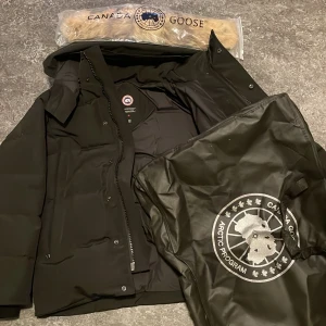 Svart dunjacka från Canada Goose S - Säljer en svart dunjacka från Canada Goose, modell Fusion i storlek S. Jackan har huva, dragkedja och knappar framtill samt klassisk Canada Goose-logga på ärmen. Yttermaterialet är slitstarkt och jackan har en avtagbar pälskant till huvan.