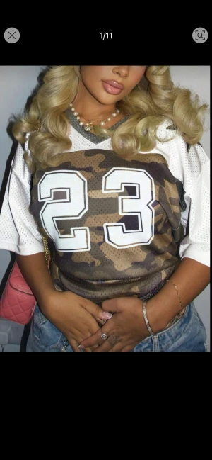 Vit och camouflage fotbollströja nr 23 - Säljer en sportig fotbollströja med nummer 23 i stort tryck på bröstet. Tröjan har vit mesh på ärmar och axlar, medan framsidan har ett snyggt camouflage-mönster i brunt och grönt. Kortärmad och tillverkad i luftig polyester.