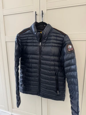 Mörkblå dunjacka Parajumpers - Snygg mörkblå dunjacka från Parajumpers med broderad patch på ärmen och orange detaljer på axlarna. Jackan har två dragkedjeförsedda fickor, hög krage och quiltad design. Perfekt för dig som vill ha en lätt och stilren jacka med coola detaljer.