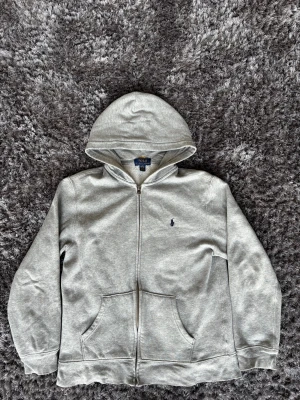 Grå hoodie från Polo Ralph Lauren - Grå hoodie från Polo Ralph Lauren med dragkedja och huva. Tröjan har ett mörkblått broderat Polo-logo på bröstet, två fickor fram och är fodrad med mjuk fleece på insidan. Perfekt för chill dagar och snygg till jeans.