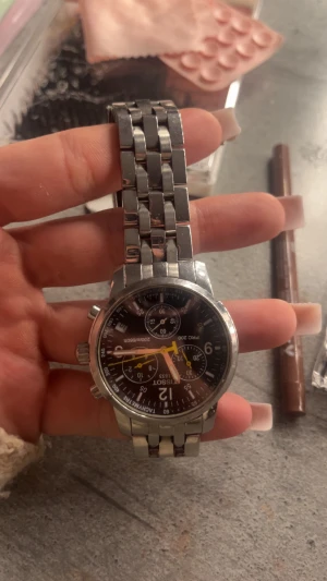 Tissot kronograf herrklocka i stål - Säljer en snygg Tissot kronograf med svart urtavla och detaljer i gult och vitt. Klockan har en rund boett och länkarmband i blankt rostfritt stål. Urtavlan har flera små visare och tachymeter-skala runt kanten. Perfekt för dig som gillar sportig och klassisk stil.