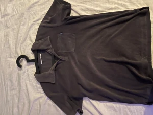 Svart pikétröja från WARP - Svart pikétröja från WARP med klassisk krage och korta ärmar. Tröjan har en bröstficka och är gjord i mjuk bomull som känns skön mot huden. Perfekt för dig som gillar enkel och stilren design.