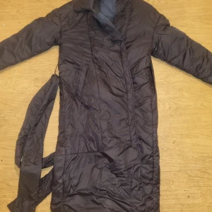 Svart lång quiltad dunjacka med bälte - Lång svart dunjacka med quiltad design och bälte i midjan. Jackan har långa ärmar, klassisk krage och två fickor framtill. Perfekt för kyliga dagar när du vill hålla dig varm och snygg.