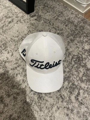 Vit Titleist keps M/L - Snygg vit keps från Titleist med svart broderad logga framtill. Klassisk böjd skärm och mesh på sidorna för extra ventilation. Perfekt för golfbanan eller som stilren accessoar. Justerbar passform och sportig look.