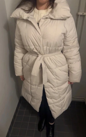 Ljusbeige lång pufferjacka med bälte - Säljer en lång, quiltad pufferjacka i ljusbeige/vitt med bred krage och knytskärp i midjan. Jackan har långa ärmar och en oversized look, perfekt för kalla vinterdagar. Snygg och trendig modell som håller dig varm hela säsongen.