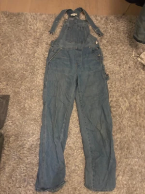 Blå jeansbyxdress från H&M, strl 34 - Säljer en klassisk blå jeansbyxdress från H&M i storlek 34. Byxdressen har justerbara axelband, stor ficka framtill och två bakfickor. 