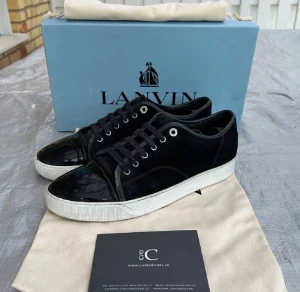 Svarta sneakers från Lanvin med lackad tå - Svarta sneakers från Lanvin med snygg kontrast mellan mocka och lackad tå. Skorna har rund tå, snörning och vit platt sula. Metallöglor och stilren design gör dem riktigt fräscha. Perfekta för dig som gillar exklusiva och trendiga sneakers.