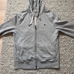 Grå zip hoodie från Morris 🙌🤩 - Grå hoodie från Morris med dragkedja och huva med vita snören. Tröjan har ett litet broderat emblem på bröstet och ribbade muddar vid ärmslut och nederkant. Perfekt för chill dagar och enkel att matcha med jeans eller joggers. Är i storlek M men passar som en S🙌🤩