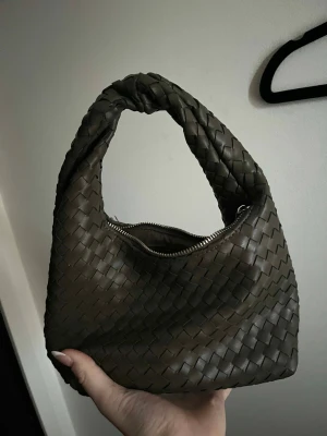 Flätad grön handväska med dragkedja - Snygg grön handväska med flätad design och praktisk dragkedja upptill. Väskan har en mjuk, rundad form och ett rejält handtag som gör den enkel att bära. Materialet är skinn och ger en lyxig känsla. Perfekt för dig som gillar stilrena accessoarer med lite edge.