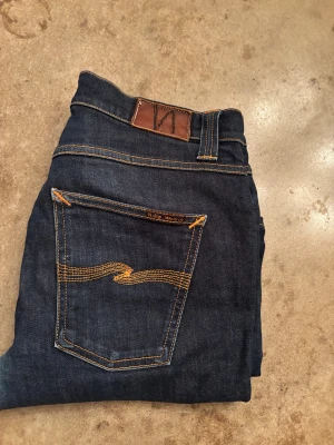 Mörkblå Nudie Jeans, storlek 31/32 - Snygga mörkblå jeans från Nudie Jeans med klassiska orange sömmar och ikonisk broderad bakficka. Modellen har normal passform och raka ben. Tillverkade i slitstarkt bomullsjeansmaterial. Perfekta för dig som gillar stilrena och tidlösa jeans.