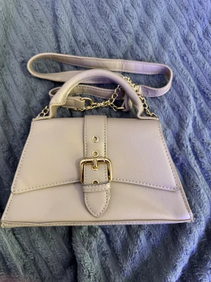 Beige handväska med guldkedja - Snygg beige handväska med guldfärgad kedja och spänne framtill. Väskan har både handtag och axelrem, vilket gör den enkel att bära på olika sätt. Stilren design med detaljer i guld och en praktisk storlek för vardagens essentials.