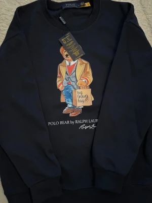 Mörkblå sweatshirt Polo Bear Ralph Lauren - Cool svart sweatshirt från Polo Ralph Lauren med ikoniska Polo Bear-trycket på bröstet. Tröjan har rund hals, ribbade muddar och mjukt material i bomull. Perfekt för dig som gillar streetstyle och vill sticka ut med en klassisk design.