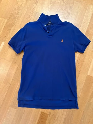 Marinblå piké från Polo Ralph Lauren - Snygg marinblå pikétröja från Polo Ralph Lauren med klassisk krage och korta ärmar. Tröjan har en orange broderad logga på bröstet och är tillverkad i mjuk bomull. Perfekt för dig som gillar stilren och sportig look.