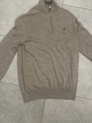 Beige stickad half zip-tröja från Morris - Snygg beige stickad tröja från Morris med half zip och hög krage. Tröjan har ett litet broderat emblem på bröstet och ribbade muddar vid ärmslut och nederkant. Perfekt för dig som gillar stilrena och klassiska plagg. Upplevs lite mindre än vanliga tröjor i S. Inga defekter!