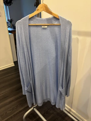 Ljusblå kofta från Isolde, storlek L - Snygg ljusblå kofta från Isolde i storlek L. Koftan är lång, har öppet framtill och två fickor. Ärmarna är långärmade med tre dekorativa knappar vid mudden. Materialet är mjukt och tunt, perfekt för lager-på-lager. Enkel och stilren design som passar till jeans eller kjol.