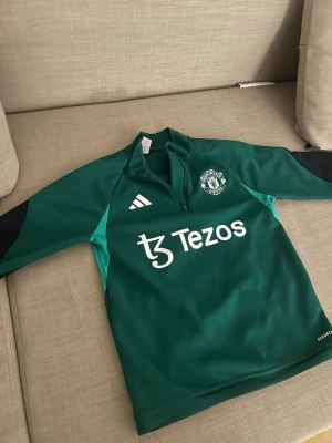 Grön Manchester United träningströja Adidas - Snygg grön träningströja från Adidas med Manchester United-logga och Tezos-tryck på bröstet. Tröjan har svart detalj på ärmarna, halv dragkedja och är tillverkad i ett funktionellt syntetmaterial. Perfekt för fotbollsträning och sportiga dagar.