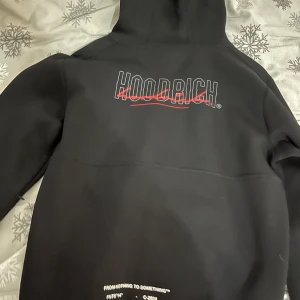 Svart hoodie från Hoodrich - Säljer en svart hoodie från Hoodrich med röd och vit logga på bröstet. Hoodien har en justerbar huva med snören och en stor ficka framtill. Perfekt för en avslappnad stil. Pris kan diskuteras!!