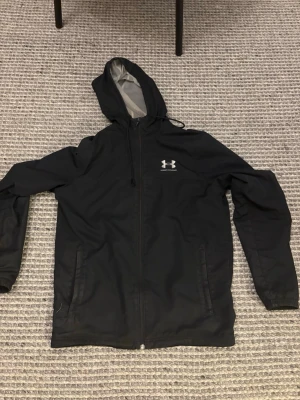 Svart vindjacka från Under Armour - Säljer en svart vindjacka från Under Armour med huva och meshfoder. Jackan har dragkedja framtill, två sidofickor och logga på