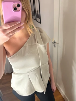 Slutsåld one shoulder topp med rosett och lappen kvar - Slutsåld one shoulder topp/blus från H&M som har lappen kvar. Är normal i storleken och har en dragkedja på sidan. På axeln sitter det en stilig och stor rosett. Blusen är åtsittande upptill och lös nedanför bysten, sluter strax över höfterna.💗💗