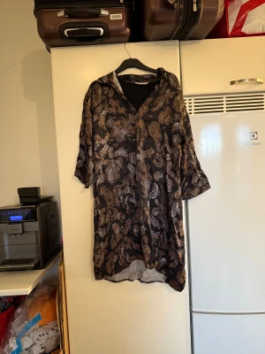 Cream klänning  - Säljer en superfin kortklänning som har en svart bottenfärg och detaljer i brunt och silver. Materialet är 100% viskos. Storlek 38. Mycket fint skick, jag hittar inga synliga anmärkningar eller defekter. 