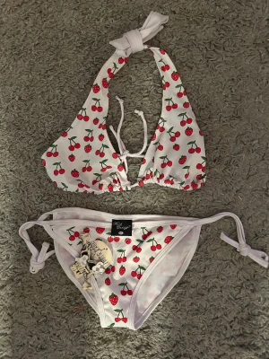 Vit bikini med körsbär/jordgubbar fr Rock Daddy. Stl M/L - Söt bikini från Rock Daddy i vit med körsbär och jordgubbar som mönster. Toppen har halterneck med knyt i nacken och framtill, och trosan har knyt i sidorna. Perfekt för dig som gillar färgglada detaljer och fruktiga prints. Helt ny.