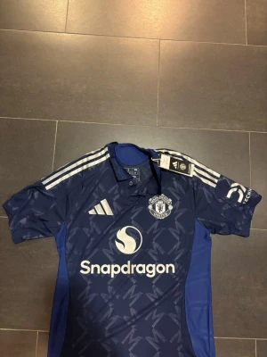 Blå Manchester United adidas fotbollsjersey  - Snygg blå Manchester United tröja från adidas i slim fit. Tröjan har vit logga, mönstrad design med M-symboler, vita ränder på axlarna och broderat klubbmärke. Materialet är lätt polyester och perfekt för fotboll eller träning. Officiell licensierad produkt med Snapdragon sponsor. Passar folk mellan 170-185. Slim fit! Tveka inte att höra av er vid frågor eller funderingar 🤗
