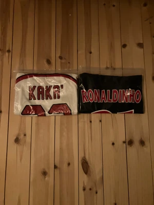 Kaká och Ronaldinho fotbollströjor - Två klassiska fotbollströjor med tryck av Kaká och Ronaldinho. En vit med röda detaljer och en svart med röda detaljer, båda med stora namntryck på ryggen. Perfekt för dig som älskar fotboll och vill hylla legenderna. Dem är inte riktiga men dem är i supper bra kvalitet