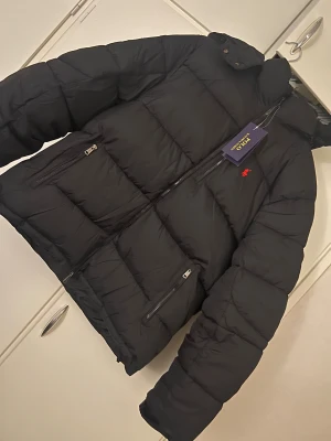 Svart pufferjacka från Polo Ralph Lauren - Säljer en svart pufferjacka från Polo Ralph Lauren med huva och dragkedja framtill. Jackan har flera praktiska fickor med dragkedjor och den klassiska röda Polo-loggan på bröstet. Perfekt för kalla dagar och riktigt snygg till streetwear-stilen.