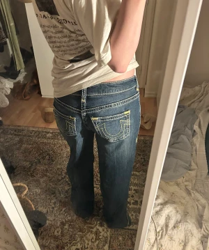True Religion Baggy blå jeans med kontrastsömmar - Säljer ett par ikoniska True Religion jeans i blå denim med tydliga vita och gula kontrastsömmar. Väldigt bra skick! Enbart smått slitna längst ned på byxbenet 