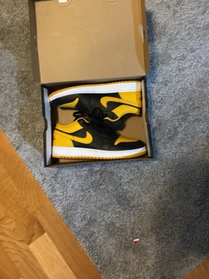 Nike Air Jordan 1 Low svart/gula  - Nike Air Jordan 1 Low sneakers i svart och gul med vit sula. Klassisk Jordan-logga på hälen och tungan, snörning och perforerad tå. Skorna har en låg siluett och är tillverkade i skinn med syntetdetaljer. Snygg kontrast mellan färgerna och ikonisk design.