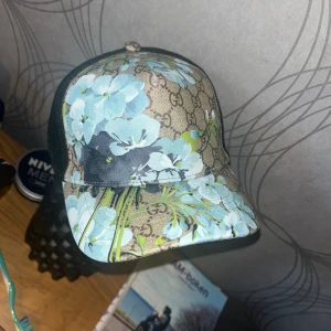 Gucci keps med blå blommor - Gucci keps med beige och svart GG-mönster, täckt av stora blå blommor och gröna detaljer. Kepsen har mesh på sidorna och justerbar baksida. Perfekt för dig som vill sticka ut med en lyxig och unik accessoar. Helt nyköpt för 650 kr! PRIS KAN DISKUTERAS!
