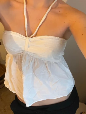 Vit halterneck bandeau topp - Säljer en söt vit bandeau topp med halterneck knyt. Toppen har en luftig och flowy passform med lätt rynk under bysten och ärmlös design. Perfekt för varma dagar och ger en fräsch sommarkänsla.