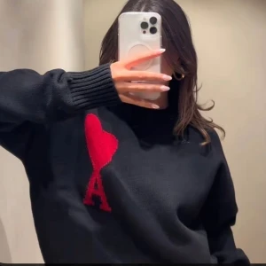 Svart stickad tröja Ami Paris med rött hjärta - Svart stickad tröja från Ami Paris med ikoniskt rött hjärta och bokstaven A på bröstet. Tröjan har långärmad design, ribbade muddar och en klassisk rund hals. 