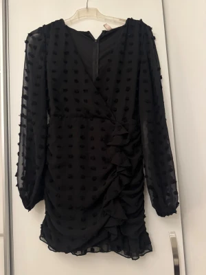 Svart prickig omlottklänning Nelly - Säljer en svart kort omlottklänning från Nelly i storlek 38. Klänningen har långa transparenta ärmar, prickigt mönster och snygga volangdetaljer framtill. Perfekt för dig som gillar trendiga och unika plagg med lite extra attityd.