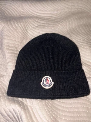 Svart Moncler mössa i ull - Svart ribbstickad mössa från Moncler med klassisk uppvikt kant och broderad logotyp framtill. Snygg och enkel design som passar till allt, tillverkad i mjuk ull för extra värme under kalla dagar.Aktiv 24/7✅ Skickas inom 24h📦🚚 Kan gå ner i hugget 😃✅ Hör av er via frågor⁉️